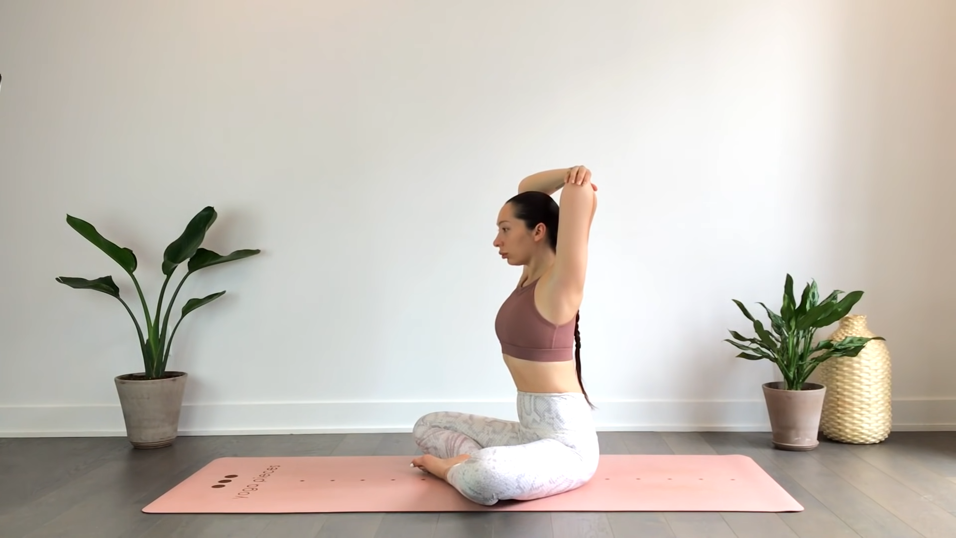 A Woman Sitting Cross-Legged on A Yoga Mat, Stretching Her Arms Overhead to Improve Flexibility in A Peaceful, Plant-Filled Room