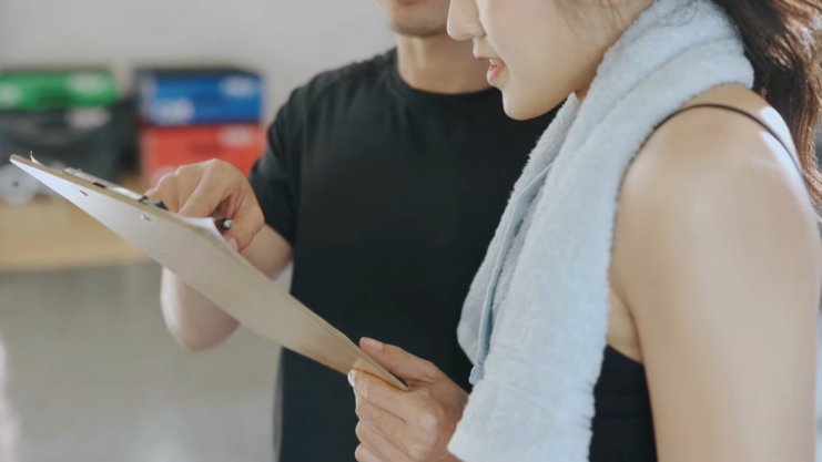 A Fitness Professional Points at A Clipboard While Discussing a Workout Plan with A Client Wearing a Towel Around Her Neck