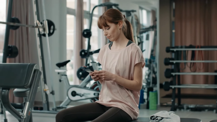 A Fitness Professional Sits Distracted on Her Phone, Appearing Disconnected While in A Gym Setting, Possibly Ignoring a Client's Needs