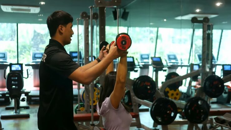 A Fitness Trainer Assists a Woman in Lifting Weights at A Gym, Guiding Her Through an Overhead Dumbbell Exercise